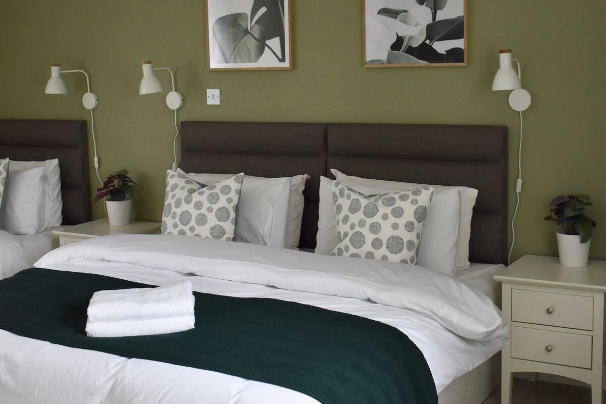 Una cama bien tendida con ropa de cama blanca y verde, dos mesitas de noche con lámparas y dos lámparas de pared.