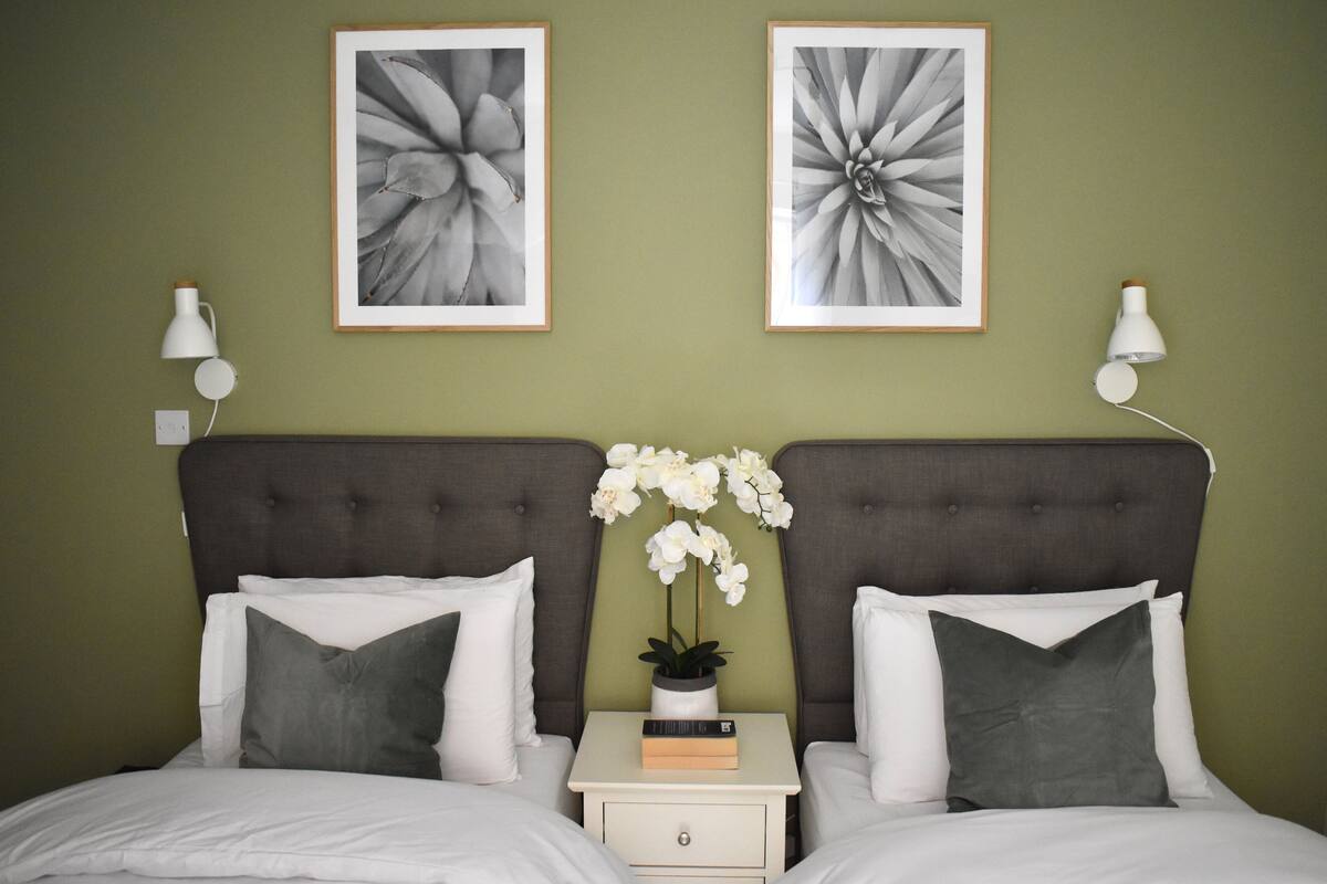 Una cama doble con sábanas blancas, dos almohadas grises, una mesita de noche con un libro y una orquídea en maceta, y dos cuadros con motivos florales en la pared.