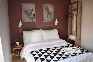 Una cama bien hecha con una colcha de estampado geométrico, una mesita de noche con una planta, y un perchero colgante.