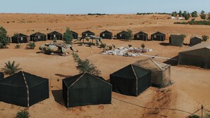 A desert camp with multiple black tents arranged in a circular pattern, surrounded by sparse vegetation and a sandy landscape. - MAMA Merzouga Camp (Rissani)