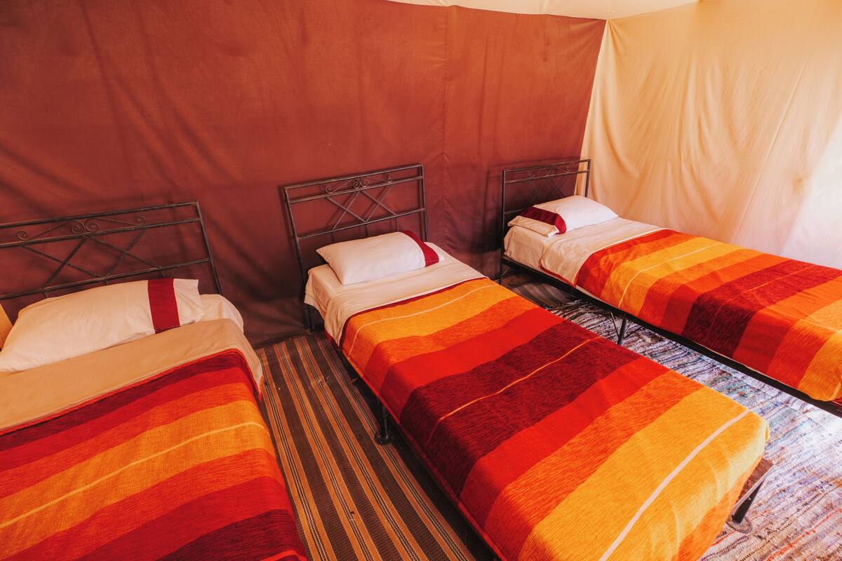 A room with two single beds, each with a striped blanket and a pillow. The beds have metal frames and are positioned against a wall with a curtain.