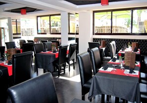 Un restaurante con sillas de cuero negro y manteles rojos, amplios ventanales con vistas al exterior y una pared acolchada de color negro.