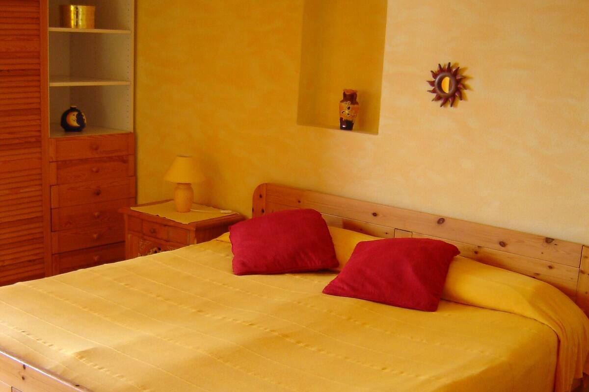 A bed with a wooden frame and a yellow bedspread, a wooden nightstand with a lamp, a wooden dresser with a mirror, and a shelf with decorative items.