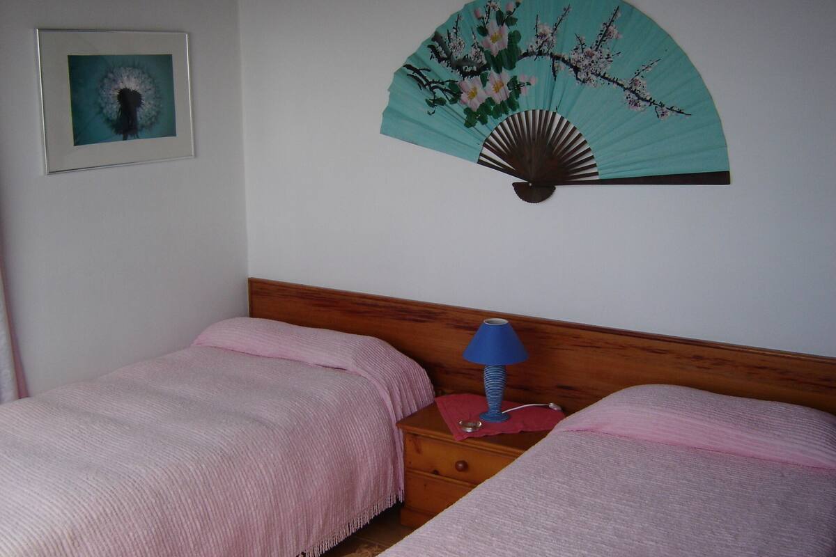 A bedroom with two beds, a nightstand with a lamp, a framed picture on the wall, and a decorative fan.