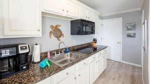 Una cocina con gabinetes blancos, encimera de granito, cafetera, lavamanos y microondas.