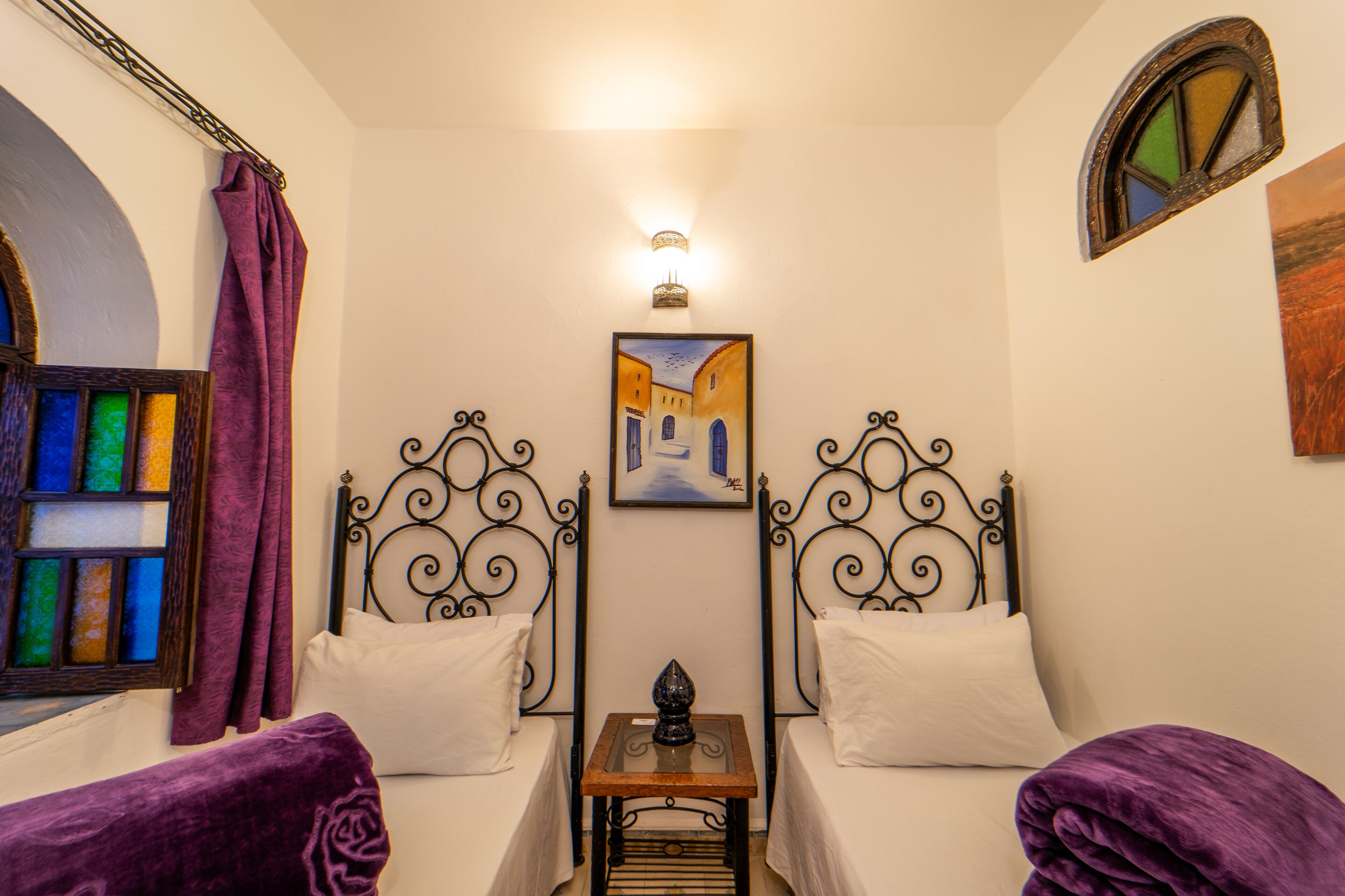 A room with two beds, a small table, a wall-mounted light, and a painting.