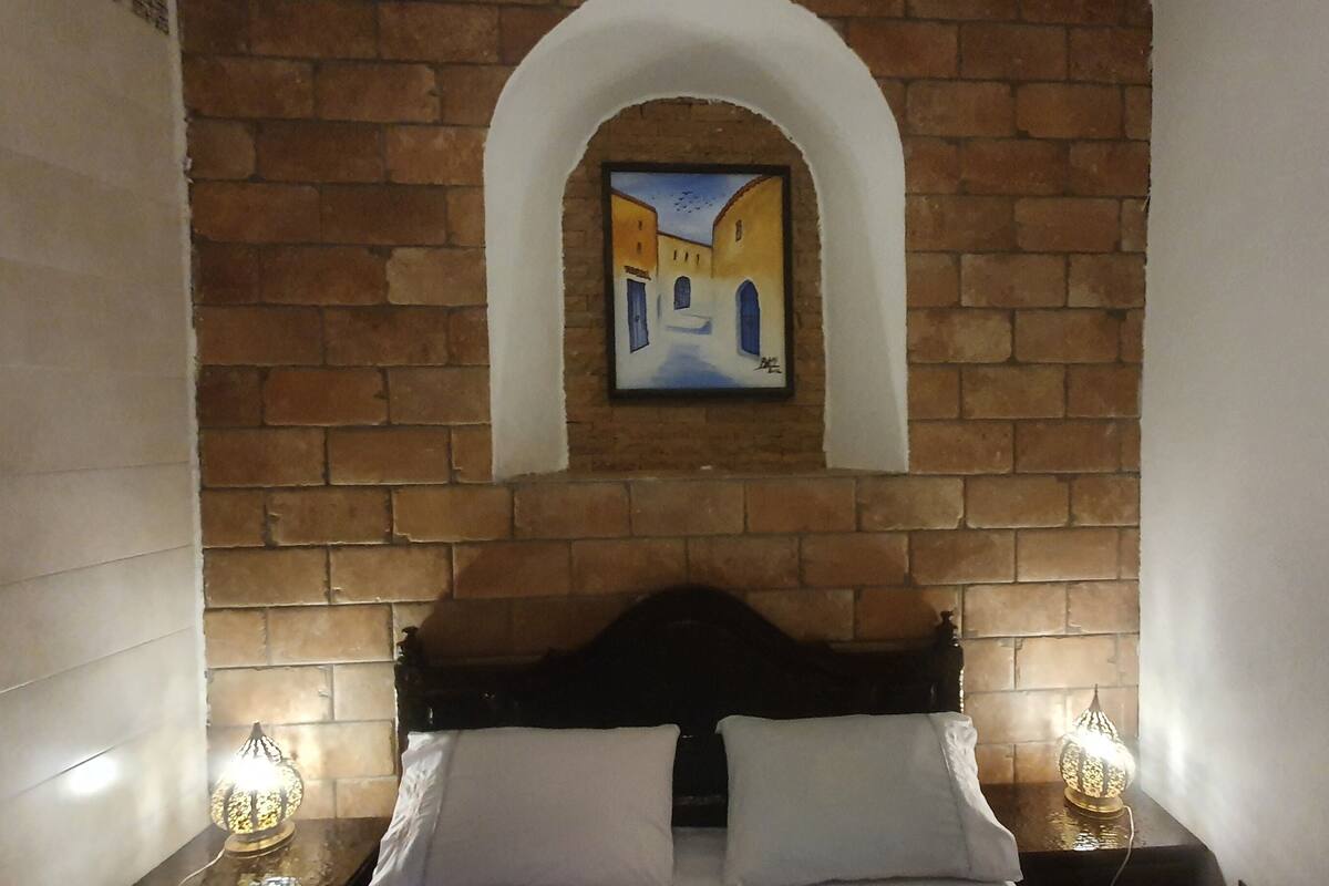Une chambre avec un mur en briques, un tableau représentant une scène de rue, un lit avec du linge de lit blanc et deux lampes de chevet.