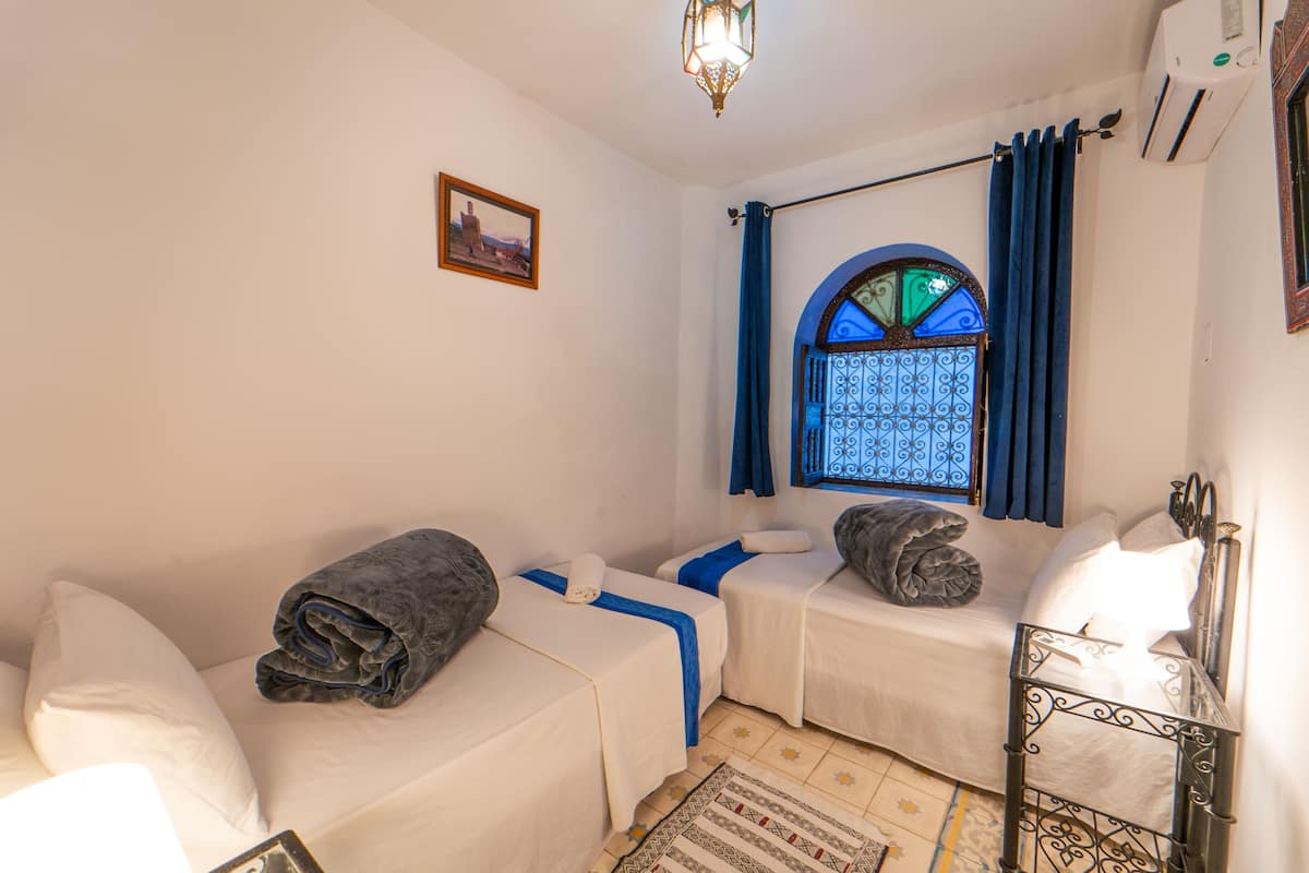 Une chambre avec deux lits, une fenêtre avec des rideaux bleus, un tableau encadré au mur et un tapis à motifs sur le sol.