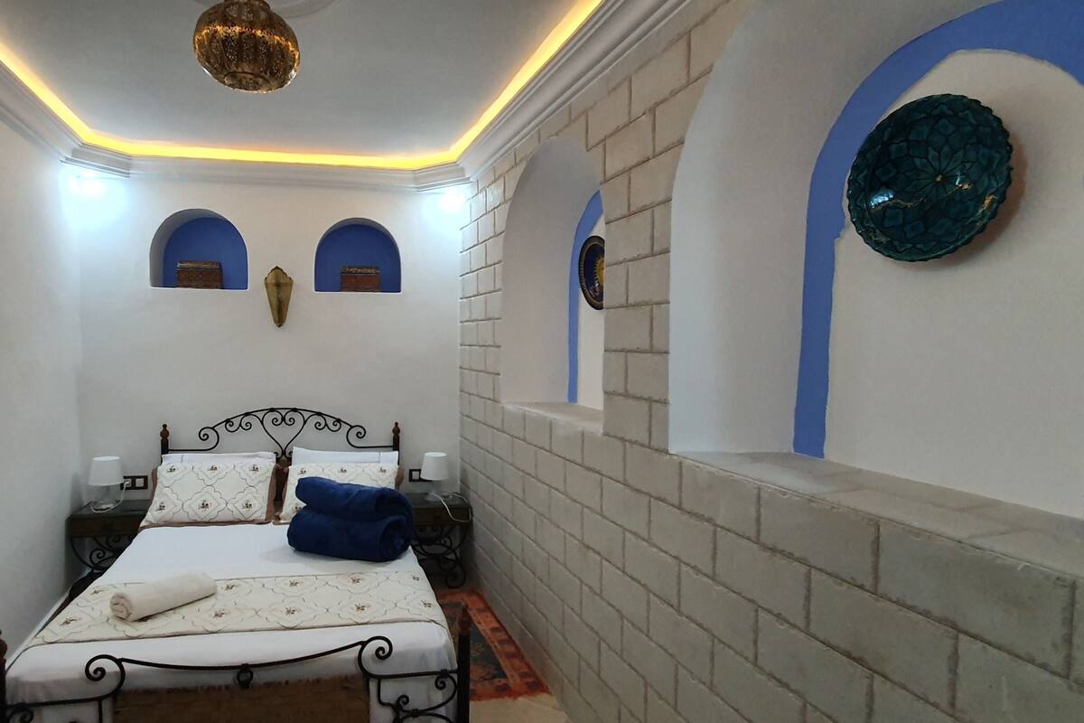 Une pièce étroite avec un lit, une petite table et un mur orné d’arches bleues.