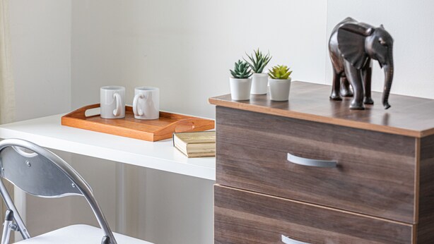 Escritorio blanco con una bandeja que contiene tres tazas blancas, un libro y dos pequeñas macetas. Hay un mueble de madera con cajones y una figurita decorativa de elefante encima.