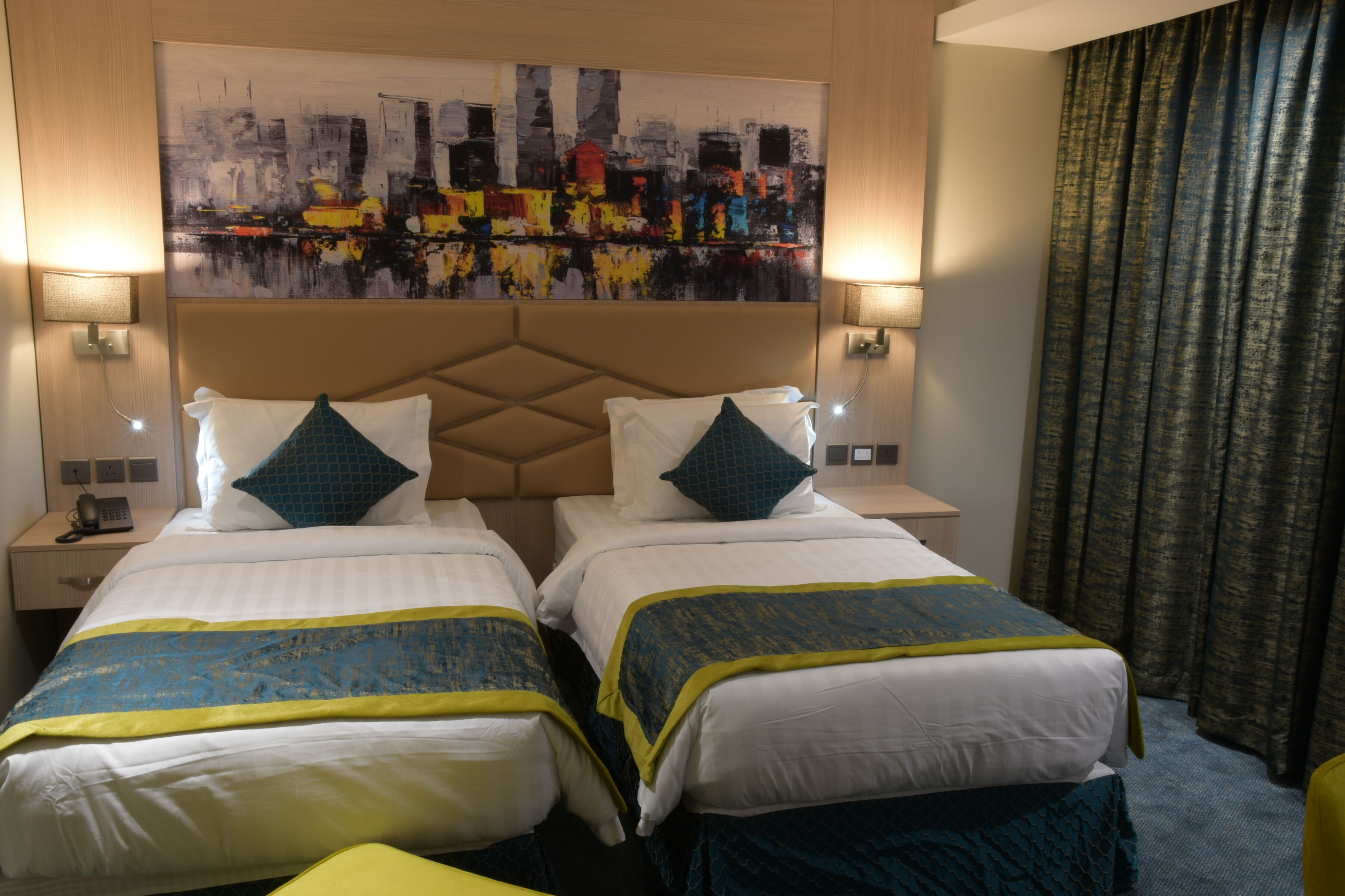 Een hotelkamer met twee bedden, elk voorzien van een hoofdeinde, witte beddengoed en blauw-gele sierkussens. Er zijn twee nachtkastjes met lampen, een groot abstract schilderij aan de muur en een raam met gordijnen.