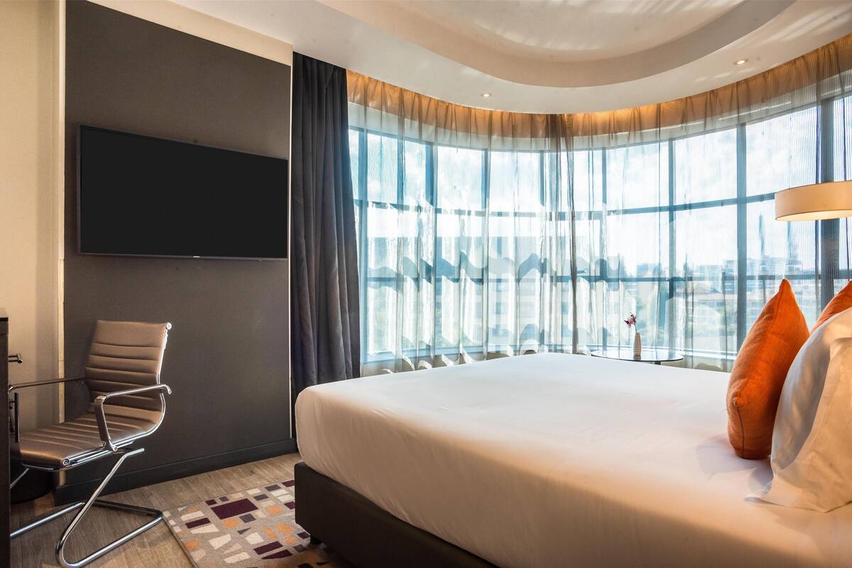 Una habitación de hotel moderna con una cama grande, un televisor de pantalla plana en la pared, una silla moderna y ventanas amplias con cortinas translúcidas.