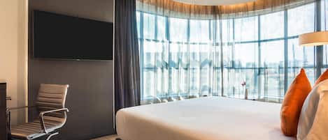 A modern hotel room with a large bed, a flat-screen TV mounted on the wall, a modern chair, and large windows with sheer curtains.