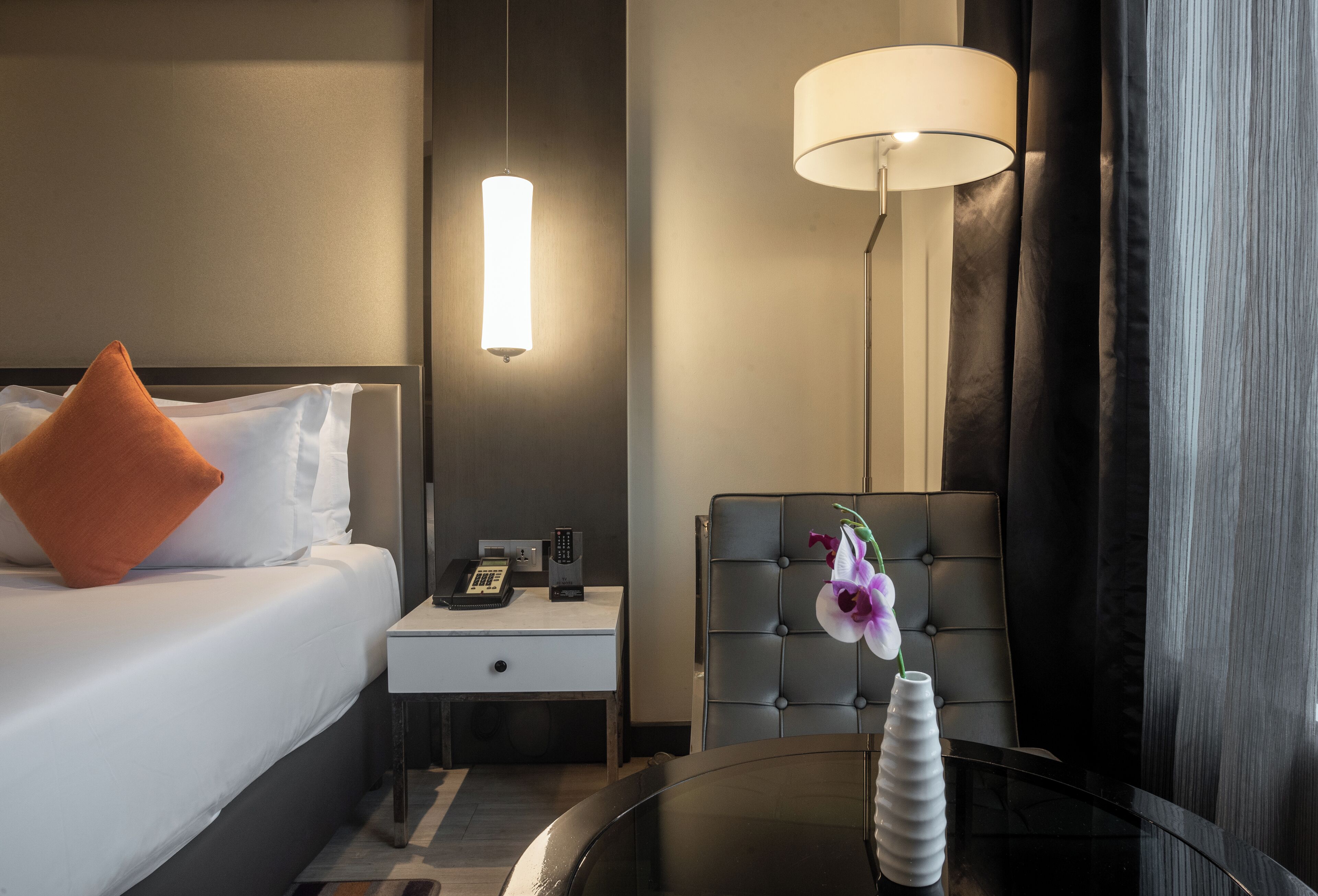 A hotel room with a bed, nightstand, lamp, chair, and a table with a vase.