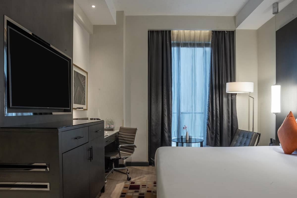 Habitación de hotel con cama, escritorio, silla, televisor, lámpara y ventana con cortinas.