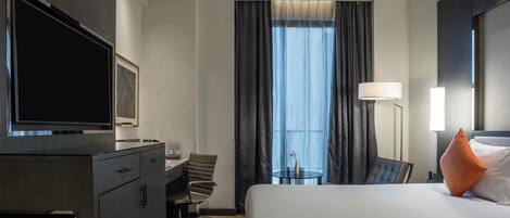 A hotel room with a bed, a desk, a chair, a television, a lamp, and a window with curtains.