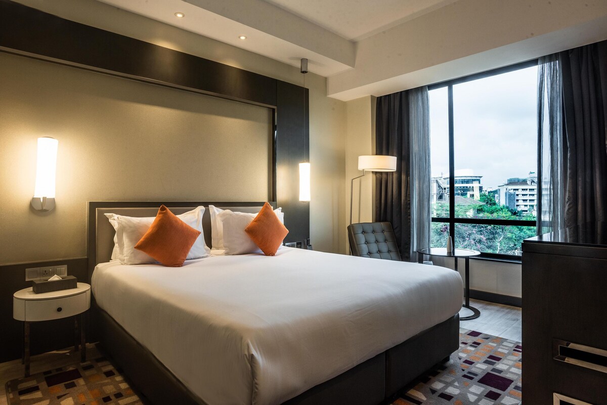 A hotel room with a large bed, two pillows, a nightstand, a chair, a small table, and a window with a view of buildings.