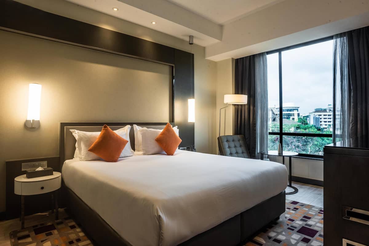 Habitación de hotel con una cama grande, dos almohadas, una mesita de noche, una silla, una pequeña mesa y vistas de la ciudad a través de amplios ventanales.