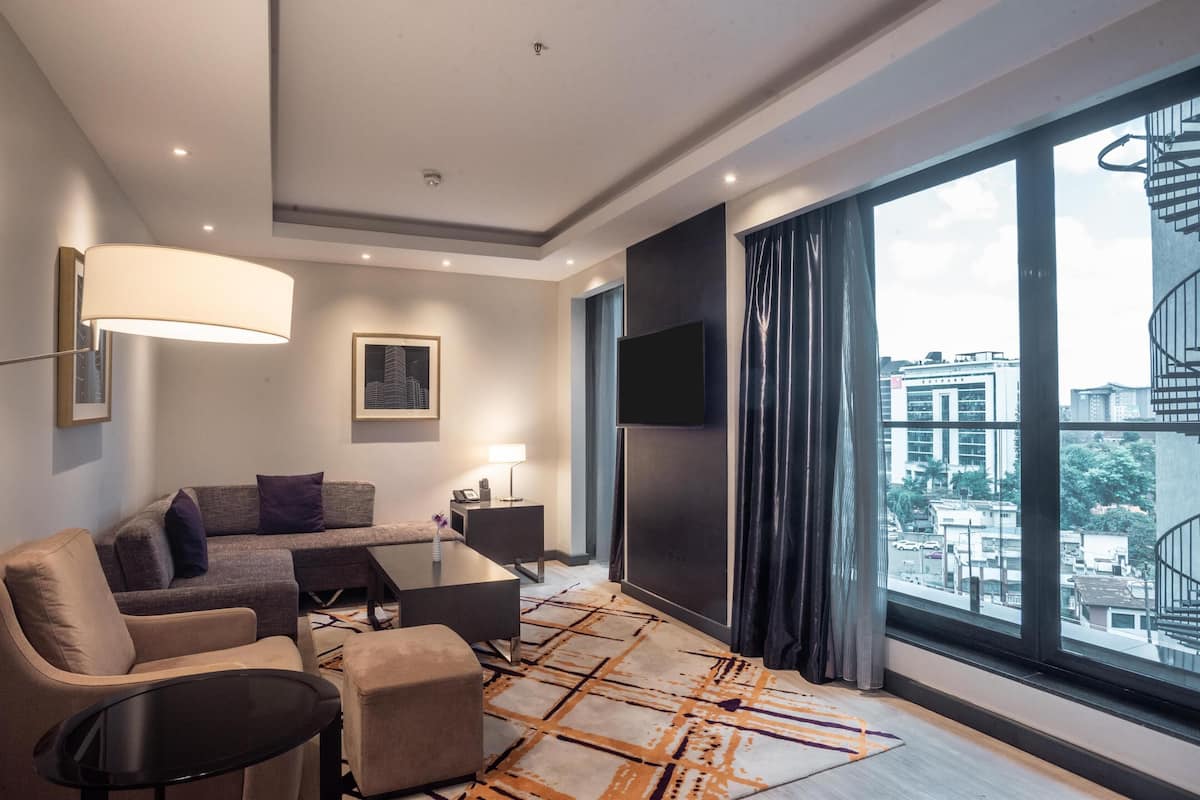 Una habitación de hotel moderna con sofá, sillón, reposapiés, una mesita, televisor y un ventanal con vistas a la ciudad.