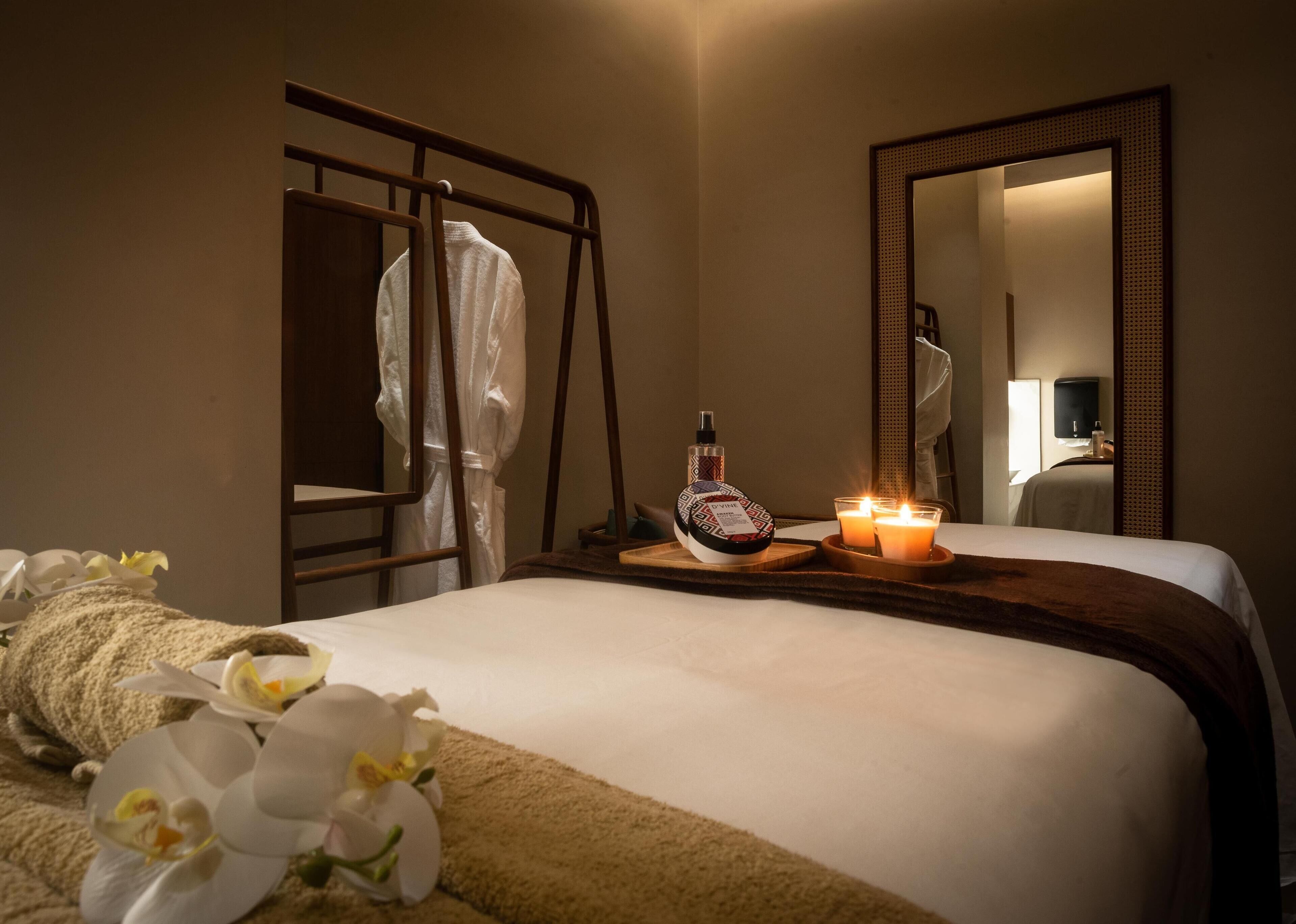 A hotel room with a bed, a bedside table with candles, a bottle, and a mirror.