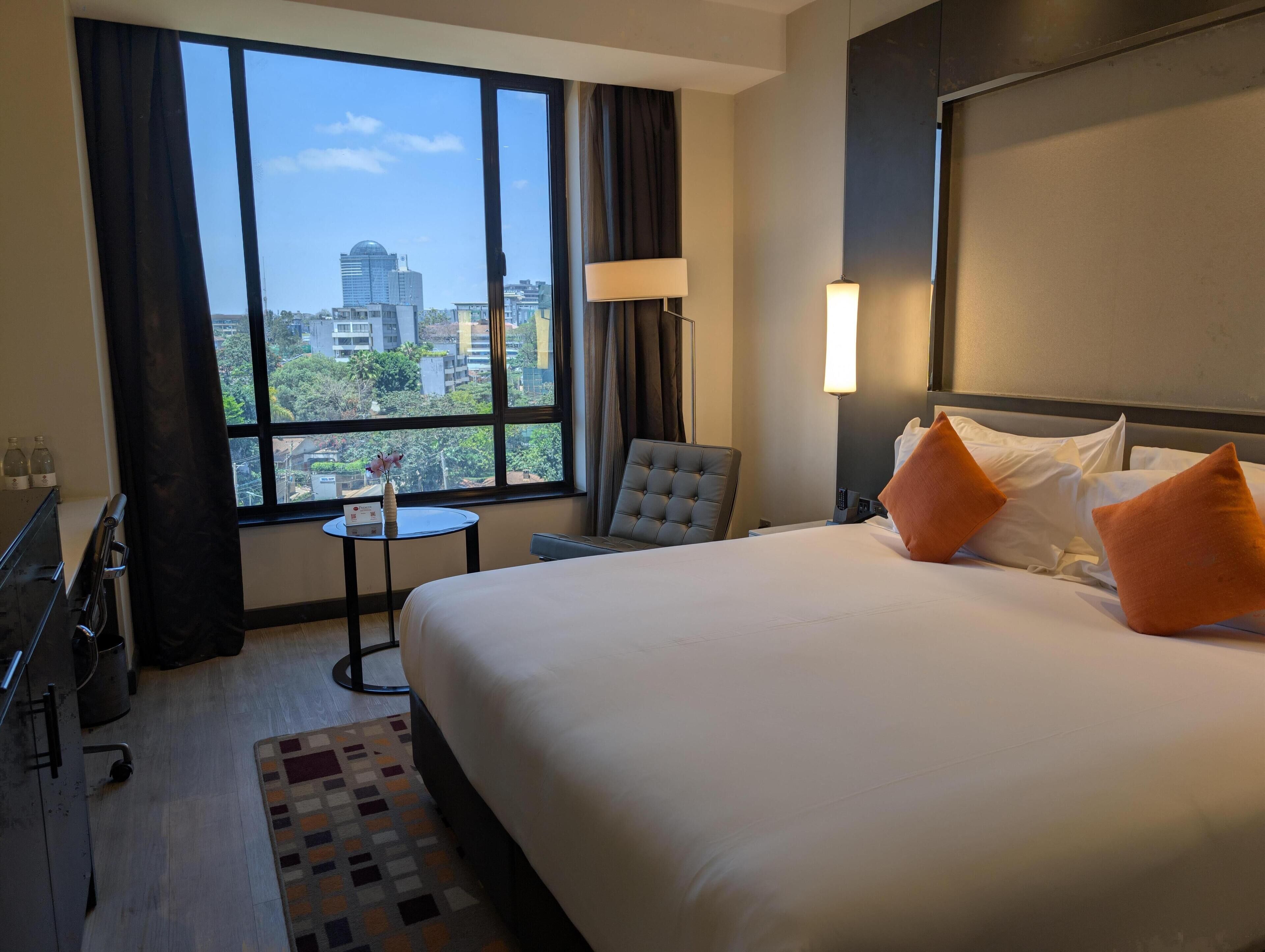 A hotel room with a large bed, a chair, a small table, and a view of the city through a large window.