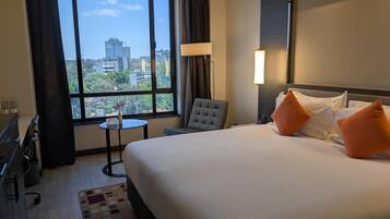 A hotel room with a large bed, a chair, a small table, and a view of the city through a large window.