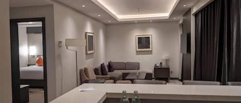 A modern hotel room with a sofa, a coffee table, a TV, and a minibar.