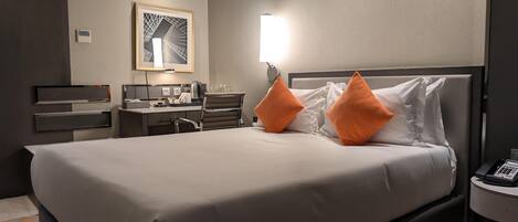 A modern hotel room with a large bed, two orange pillows, a desk with a chair, a coffee maker, and a framed artwork on the wall.