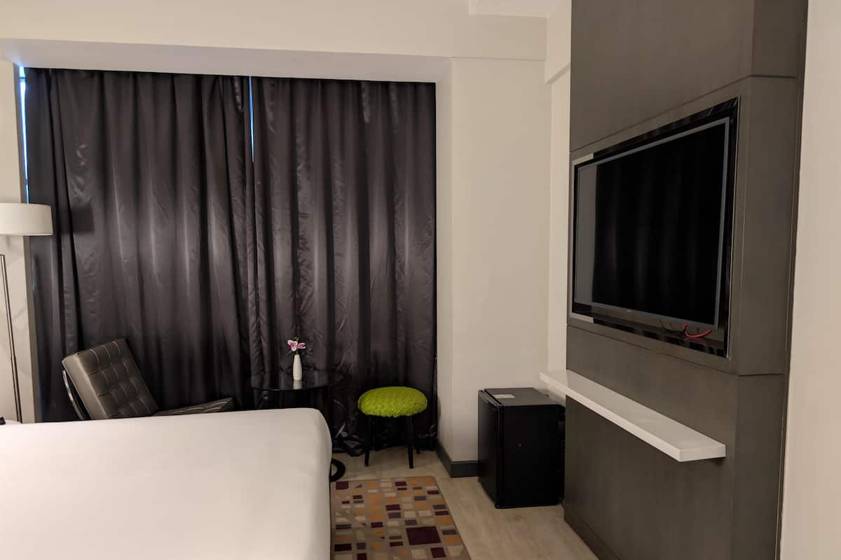 Habitación de hotel con cama, televisor montado en la pared, una mesita con un florero, una silla y alfombra estampada.
