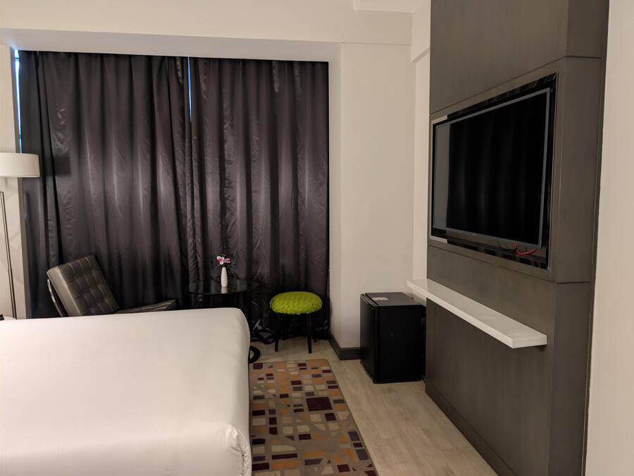 A hotel room with a bed, a TV mounted on the wall, a small table with a vase, a chair, and a patterned rug.