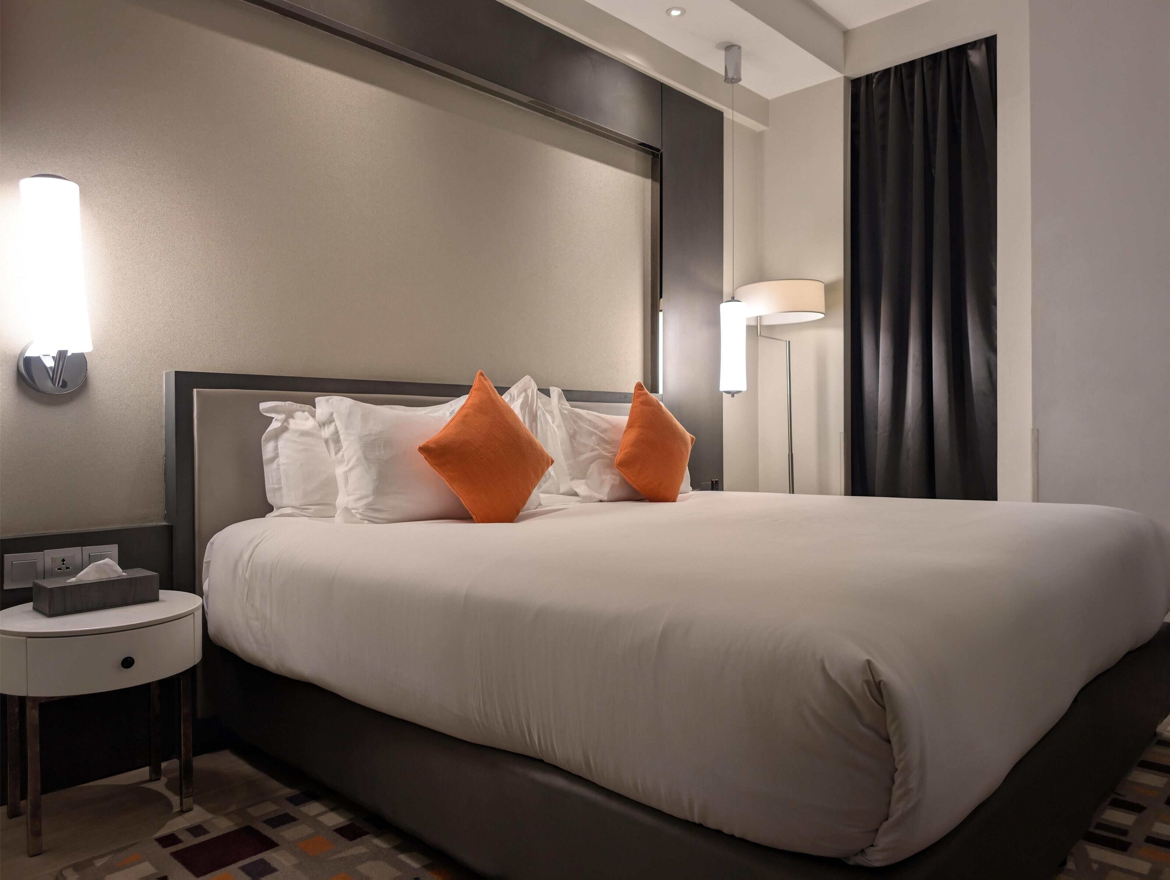 Una habitación de hotel con una cama grande, ropa de cama blanca, dos almohadas naranjas, una mesita de noche con una lámpara y un mueble auxiliar con una caja de pañuelos.
