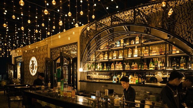 A bar area with a curved shelving unit displaying bottles and glasses, a counter with bar tools, and hanging lights.