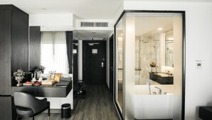 A modern hotel room with a dark wood floor, a black upholstered chair, a small trash bin, a glass-enclosed shower area, and a white sink.