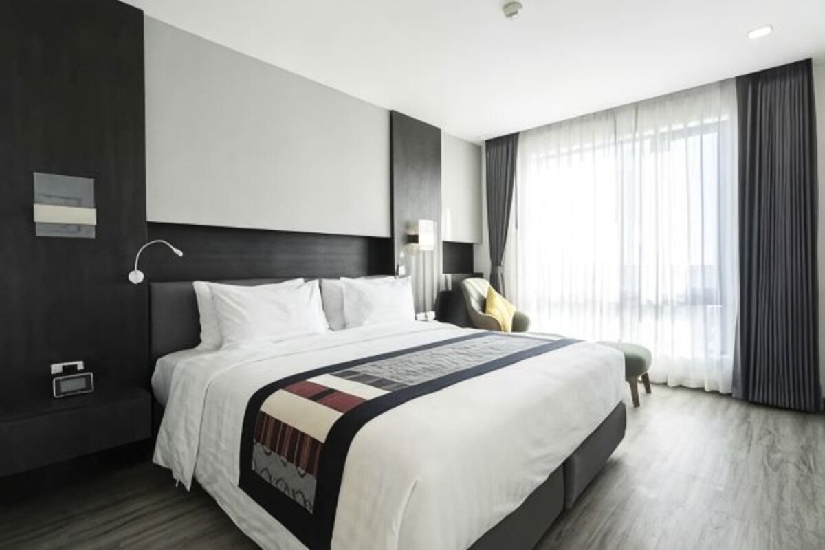 Habitación de hotel moderna con una cama grande, una mesita de noche, una silla y un ventanal con cortinas.
