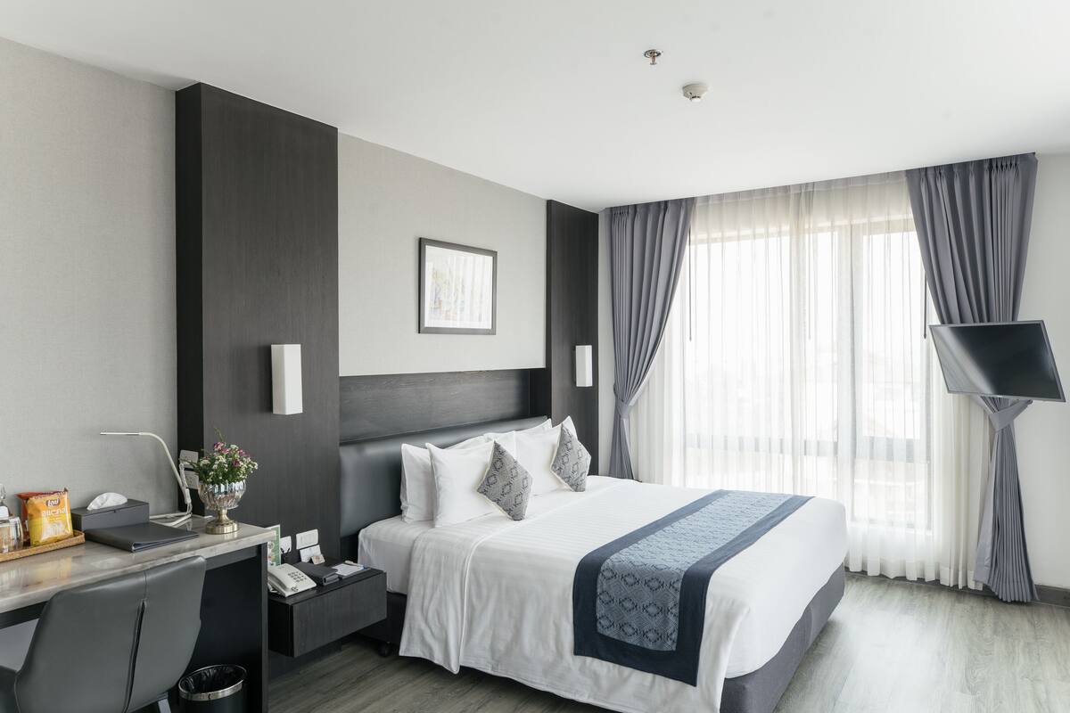 A modern hotel room with a large bed, a desk with a chair, a flat-screen TV, and a window with curtains.