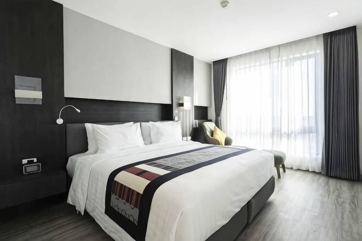 A modern hotel room with a large bed, a bedside table, a chair, and a window with curtains.