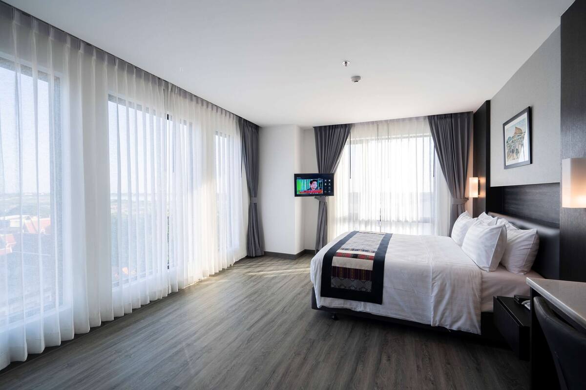 A hotel room with a large bed, a flat-screen TV, and a view of the city through sheer curtains.