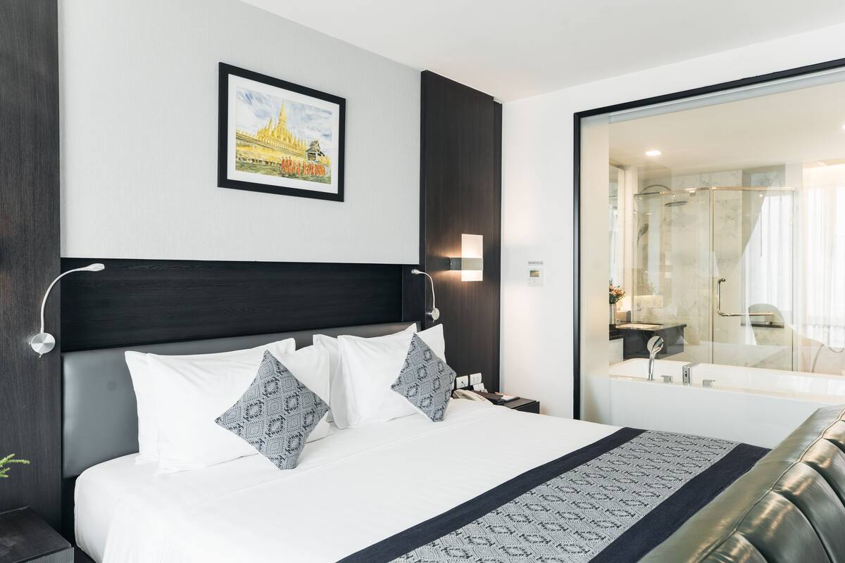 A modern hotel room with a large bed, a nightstand, a framed picture on the wall, and a view of the bathroom.