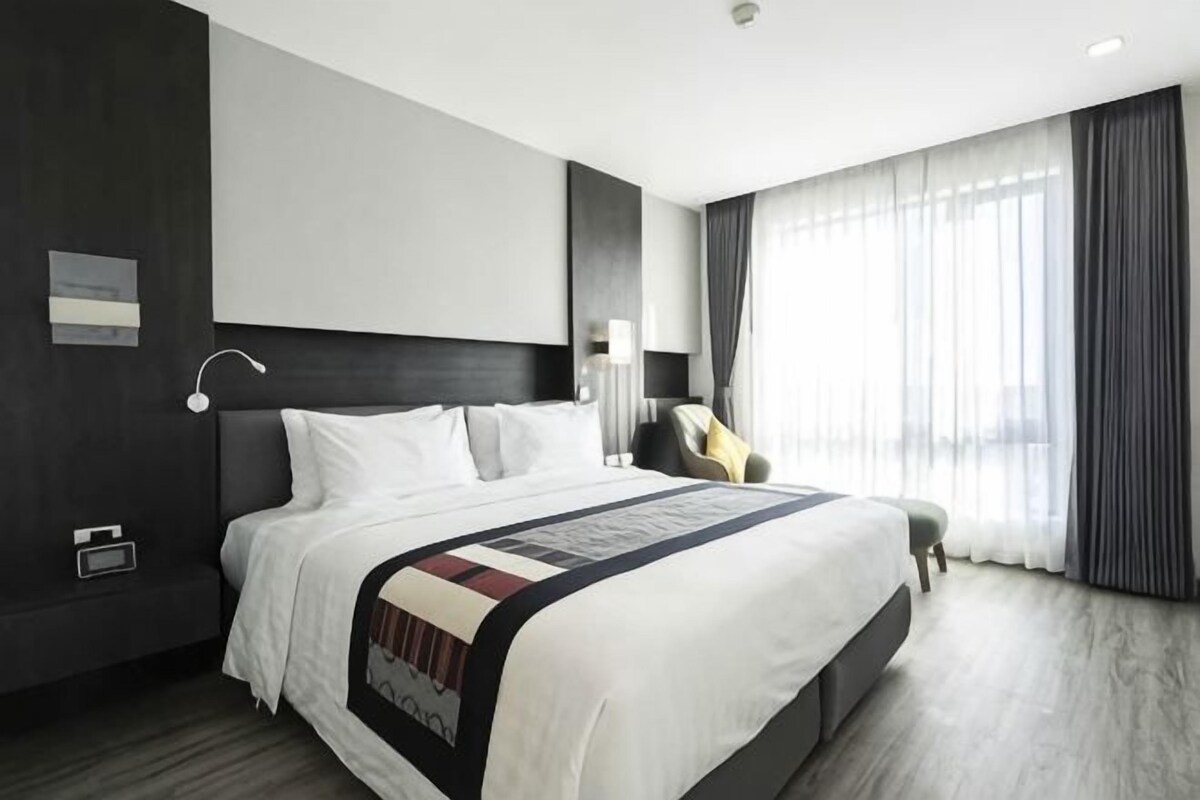 Habitación de hotel moderna con una cama grande, una mesita de noche, una silla y un ventanal con cortinas.