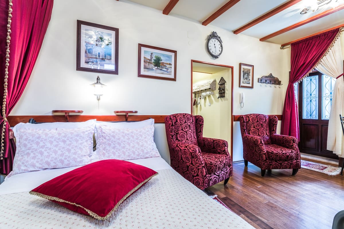 Een kamer met een bed, twee rode fauteuils met een patroon, een houten plafond en een wandklok.
