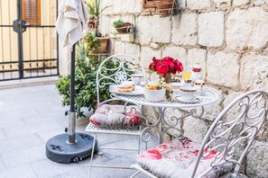 A metal table with a white teapot, cups, and pastries. There are chairs with floral cushions and a potted plant in the background. - Emperor's Suites (Split)