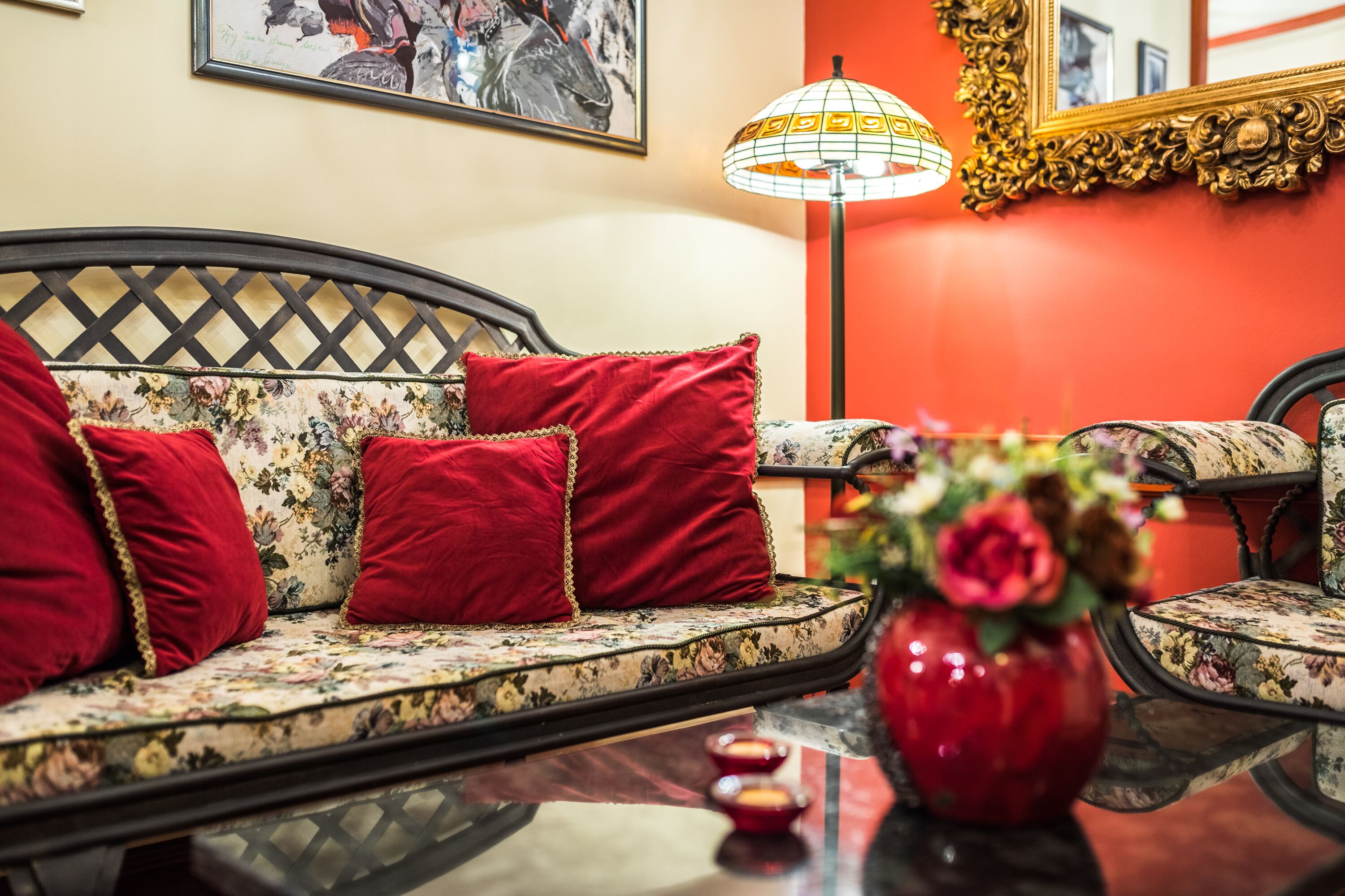 A sofa with floral upholstery and red cushions, a red vase with flowers, and a lamp with a patterned shade.