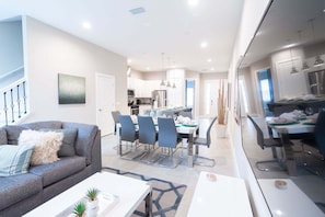A modern living room with a sofa, coffee table, dining area with chairs, and a kitchen in the background.