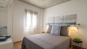 Une chambre à coucher avec un lit, une fenêtre avec des rideaux, une lampe fixée au mur et une petite plante posée sur une table d’appoint.