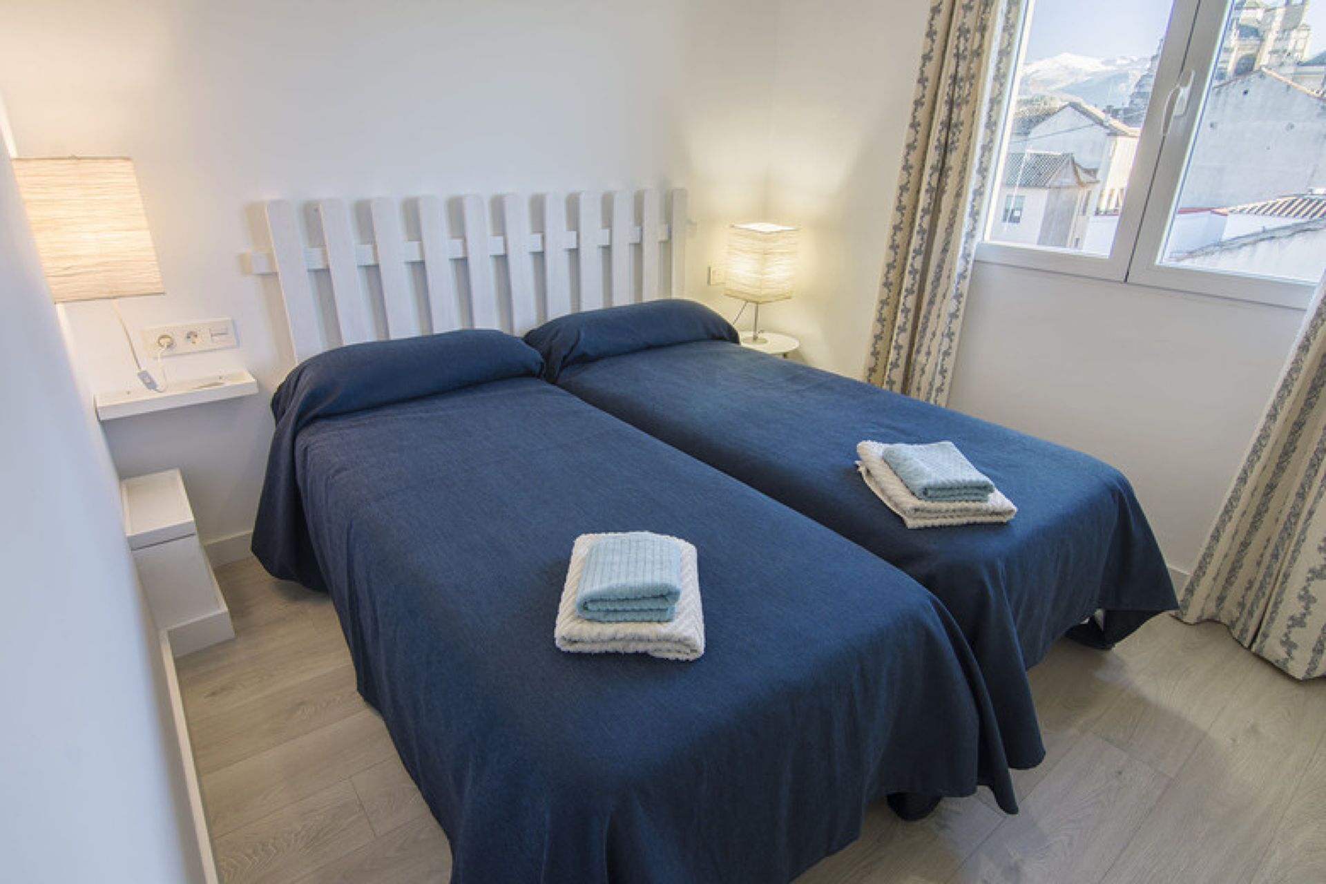A double room with two beds, a bedside table, a lamp, and a window with curtains.