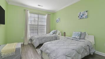 Un dormitorio con dos camas, una ventana con persianas, una mesita de noche con lámpara y un televisor montado en la pared.