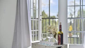 A wooden tray with a wine bottle and two glasses on a table by a window with white curtains.