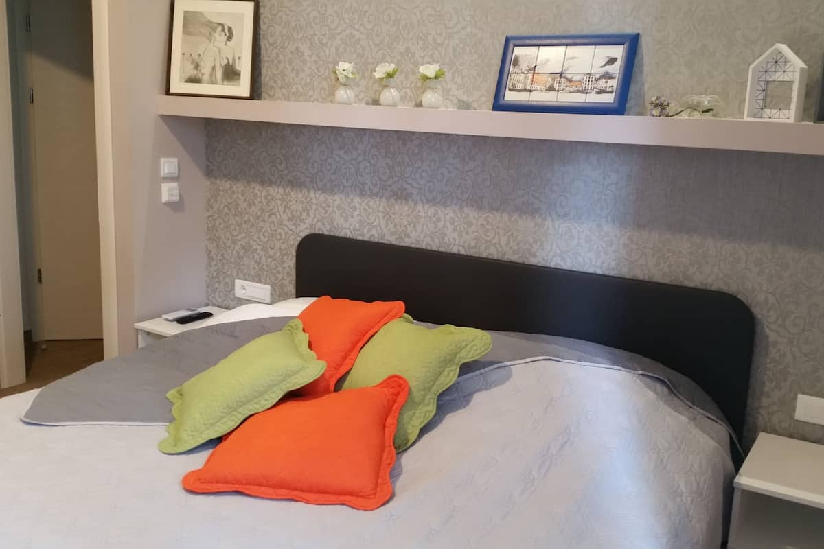 Una cama bien tendida con almohadas coloridas, una mesita de noche con un teléfono y un control remoto, y una pared con obras de arte enmarcadas y adornos decorativos.
