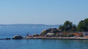 Zona costera con un puerto deportivo, barcos y una playa con sombrillas y gente.