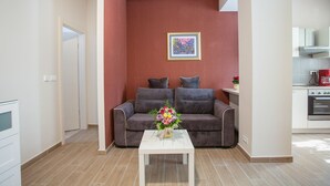 En moderne stue med en grå sofa, et hvidt sofabord med blomsterarrangement, samt et køkkenområde med en rød elkedel.